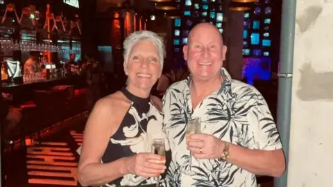 Trevor Davies A woman wearing a summer dress which is black with a white flowery pattern and a man wearing a white shirt with black flowers on. They are holding glasses with drink in. 