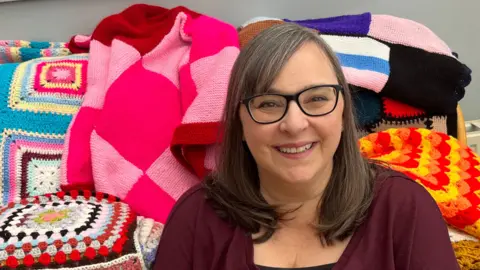Kathleen Dabbs has long dark hair parted on her head. She has black-framed glasses on and a purple and black top on. She is on a sofa surrounded by lots of colourful blankets.