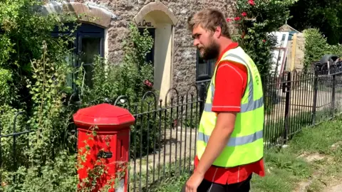 A man with dark blonde hair and beard stands sideways on to the camera looking at a red postbox. He is wearing a red short-sleeved Post Office top covered by a neon yellow tabard. The postbox stands in front of black iron railings, shrubs and a stone building with low arched windows.