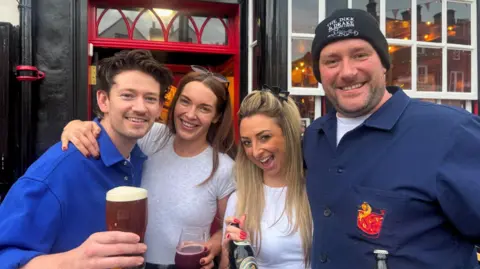 Two men and two women stand closely together outside a traditional British pub, smiling and enjoying drinks. The pub has a black exterior with red-framed windows. Two individuals hold pints of beer, one holds a glass of red wine, and another holds a bottled drink.