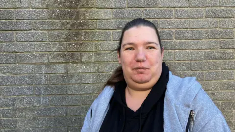 A woman wearing a black top and a grey zip up hoodie looking into a camera. She is standing in front of a grey brick wall.