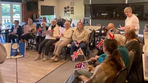 Moorlands Home Link A group of older people, some wearing paper hats and holding Union Jack flags, are sat in a canteen. They appear to be taking part in a group activity such as singing.
