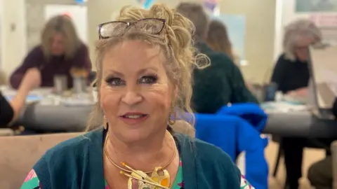 A woman in colourful clothing and a large necklace with a pendant in the shape of a palette and paintbrushes. Multiple people are painting at a table in the background.
