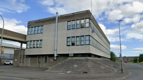 Google Street view image of Washington Police station, on the corner of two streets. It's a large, brutalist, grey building.