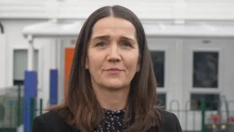 Jamie Niblock/BBC Academy trust chief Jo Coton in front of a white school building. She has long brown hair and is pouting while wearing a black blazer and navy and white top.