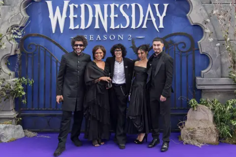 Getty Images Moosa Mostafa, centre, is flanked to the right by mum Rima, and dad Hani, who is wearing dark glasses. To his left is sister Roo and cousin Omar. All wear black clothes and are standing in front of a purple backdrop with Wednesday, Season 2 in white lettering