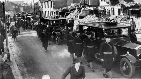 Getty Images A black and white image of funeral cars drive slowly through a bomb-damaged street in Portsmouth, accompanied by an escort of British soldiers and sailors. Twenty-five victims of an air raid are to be buried in a common grave. 