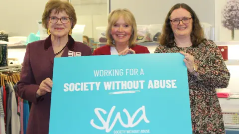 Three women stand in a charity shop holding a large blue sign saying 'Working for a society without abuse'