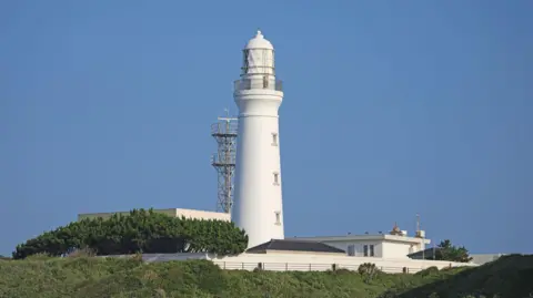 Getty Images A white lighthouse surrounded by other low buildings and trees juts out into a blue sky