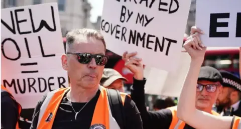 Dale Vince stands in the middle of a protest with white protest posters being held around him. He is wearing a florescent vest, sunglasses and a black t-shirt.  