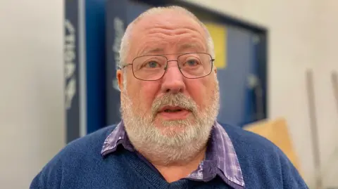 John Lemon of East Belfast Men's Shed looks behind the camera. He is standing in front of a white wall with a blue object behind him. John has short, white hair and is wearing a pair of silver framed glasses, a blue jumper and a purple plaid shirt.