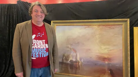 Scott O'Hara stands smiling beside a Turner painting. He is wearing a light brown jacket and a red T-shirt with red and blue writing on it