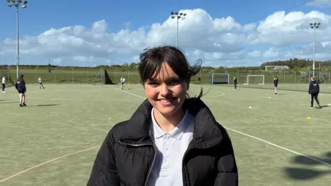 Molly, a pupil at Cockermouth School, stands in front of the astro turf cricket pitch wearing a white polo shirt and black jacket.