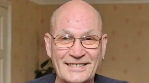 SWNS David Newton: A man is smiling at the camera in an image from 2016. He is bald, has some stubble and wears thin framed glasses. He is wearing a navy jumper, with a grey top underneath a has a hooped earring in his left ear. He is inside a room with rust colour wallpaper that has red diamond shaped patterns. There is a white door behind him to his left and a green plant.