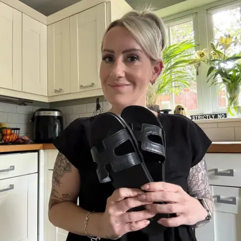 BBC Emma Neil with blonde hair holding her black sandals in her kitchen