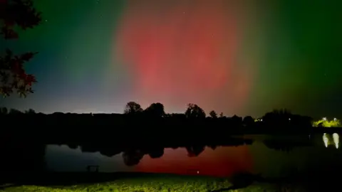 JA/BBC News NI Weather Watcher Northern Lights in the sky above a dark body of water. There are streaks of blue, green and red in the sky. 