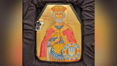 The icon of St Volodymyr The Great is painted on a piece of damaged body armour. There is a large bullet hole on the left hand side of the plate, which is now painted with a golden background and in a padded box for protection.