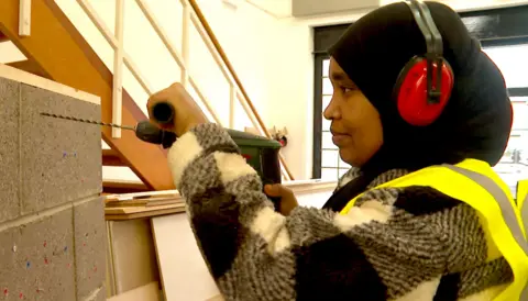 Samira is wearing a black and white checked shirt, a hi-vis yellow vest and red ear protectors. She is holding a drill and drilling into some grey masonary in front of her..