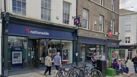 LDRS A Nationwide bank branch sits along a row of brick and painted shopfronts on a high street. The dark blue Nationwide sign hangs above large windows and the entrance, with an ATM visible beside the door. Several people walk past the shop, while others stand nearby. A row of bicycles is parked in front of the building, and additional shops and signage continue along the street.