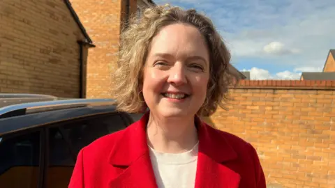 Anna Sabine smiling at the camera. She has short blonde hair and is wearing a red coat with a cream top underneath. She is standing in front of brick walls on a cloudy day.