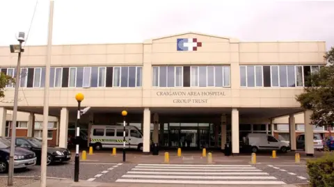 Pacemaker A big off-white hospital facade with a medical crest below the ridge. Parked out the front are ambulances and there is a zebra crossing in foreground, 