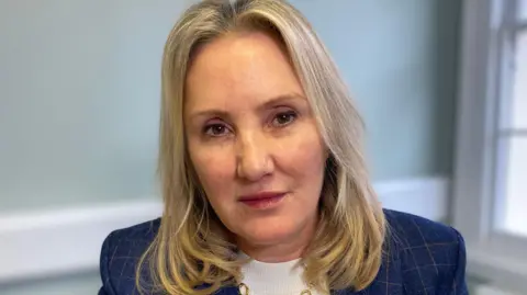 Gosport MP Dame Caroline Dinenage is pictured looking at the camera - she has a blue checked jacket and white top and shoulder length blonde hair.