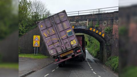 A HGV vehicle titled at a 45 degree angle and lodged underneath a bridge