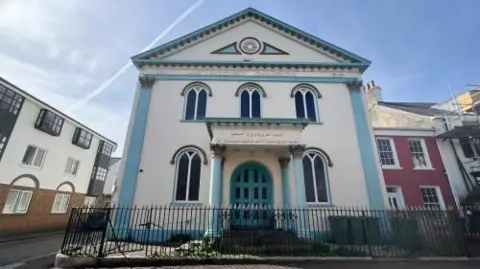 The Jersey Islamic Centre is a former Methodist church and is painted white and blue. Next to it are two residential homes, one small pink one and one larger white one with green shutters and a hedge in front of it
