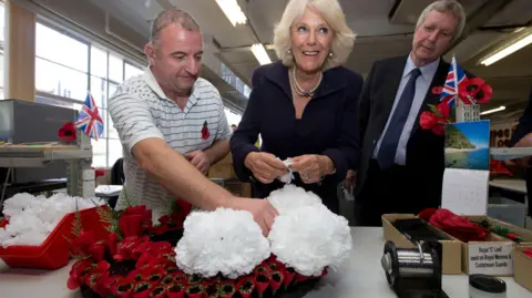 Man in striped shirt holds white flowers on top of red poppy wreath as woman with pearls smiles and prepares to add flowers