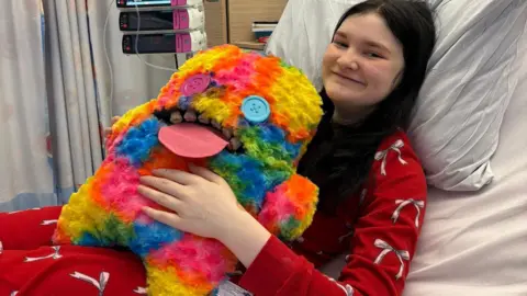 Alexa Perkins has long black hair and wears red pyjama top and bottoms with pink bows. She is laying on a hospital bed with a large multi-coloured cuddly toy. She is smiling.
