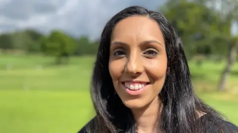 Gurj Bowbanks, who has black hair, is pictured smiling at the camera with the golf course blurred in the background.