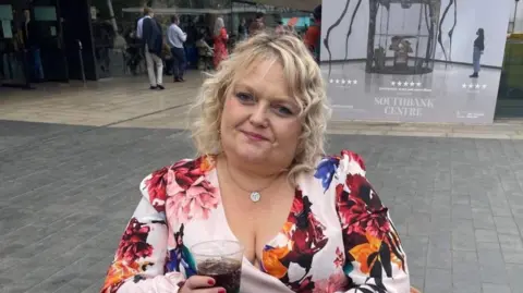 Brain Tumour Research A woman with blonde hair and wearing a dress with a floral pattern is holding a drink.