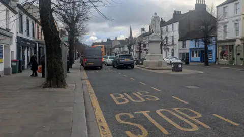 A general view of the bus stop on Cockermouth Main Street. The road is busy with cars and vans and there are a few people standing at the stop.