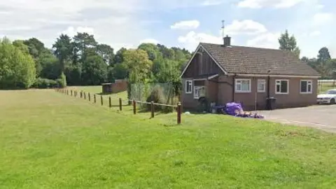 A brick building with a slate roof with a field to the side. There is a white car parked by the side of the building. 