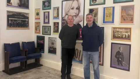 MNH Matthew Richardson and Seamus Shea, who both have short greying hair and are wearing jeans and jumpers, standing in front of a wall covered in pictures of performers and posters of old shows.