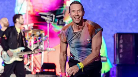 Chris Martin of Coldplay on stage in front of a colourful backdrop. He is smiling at the crowd