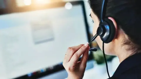 Getty Images Shot from behind of a woman with dark hair wearing a telephone headset. She has her left hand touching the microphone arm. There is a screen in front of her.