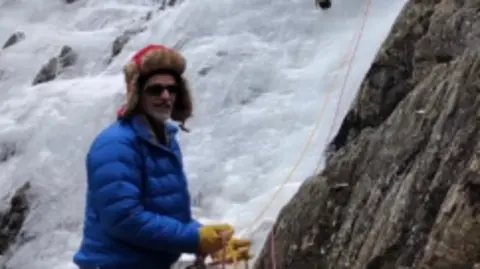 Police Scotland Man wearing a red helmet, blue puffer jacket and yellow gloves climbing an icy rock face, secured by ropes.