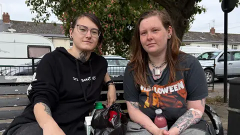 Two women in their 20s, both with tattoos, sit on a bench with a tree behind them.