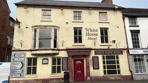 A three-storey 18th-century inn. It's painted a faded yellow, with a dark red front door. The nine windows are boarded up, and the door is nailed shut. A sign above the right-hand first-floor window reads 'White Horse Hotel', which is in yellow font on a black sign. To the left of the building is a road sign, directing cars to Ellesmere, Whitchurch, Prees, the train station and the swimming pool.