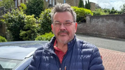 Charlie Stubbs / BBC There is a man in a dark blue puffer jacket, a red polo and round-framed glasses. He has brown hair which is wind-swept up and a brown and grey stubbly beard. He is stood with his car and greenery over his right shoulder.