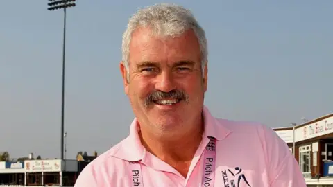 The Minor Counties Cricket FestivaL A head and shoulders image of Don Topley. He is wearing a pink polo shirt and smiling directly into the camera. He is standing in what appears to be the Essex County Cricket ground. 