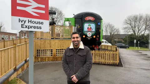 Baasit Siddiqui - from Gogglebox - stood next to a train sign which says "Ivy House Express" behind him is the front of the converted train carriage, which is black at the front and includes an image of a driver.