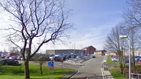 Google A view of the HMP Lowdham Grange in Nottinghamshire carpark showing a road and cards in the foreground and buildings in the background. 