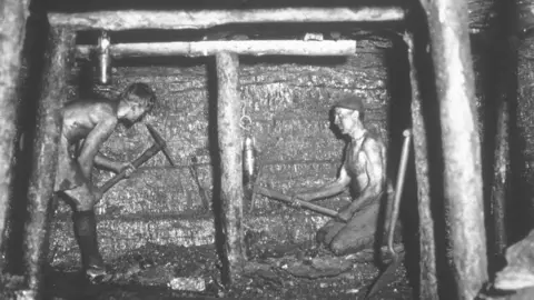 Getty Images Two topless men work in a tiny dark crawl space underground. 