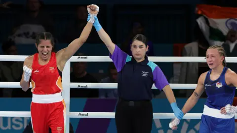 PA An official raises the arm of celebrating Sakshi Chaudhary in a red India uniform to signal victory, while Ruby White in a blue England uniform stands on the opposite side of the ring visibly upset.