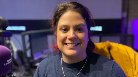 Regan Harris smiling in a radio studio with a purple Radio Lincolnshire microphone to the left of the picture. She is wearing a blue jumper and a silver necklace.