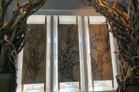Either side of the window are intertwined tree branches. The windows have brown blinds and the fabric is decorated with illustrations of trees without leaves.