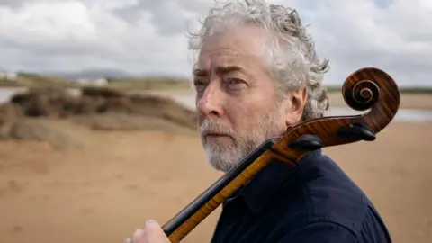 Martin has shaggy grey hair and a short white beard. Ge is on a beach the neck of a cello is propped against his shoulder.