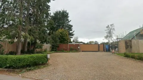 Google Images The image shows a piece of land opposite a farm. The sky is grey and there are trees growing on the left hand side. A house can be seen in construction on the right and a Union Jack can also be seen on top of the house.